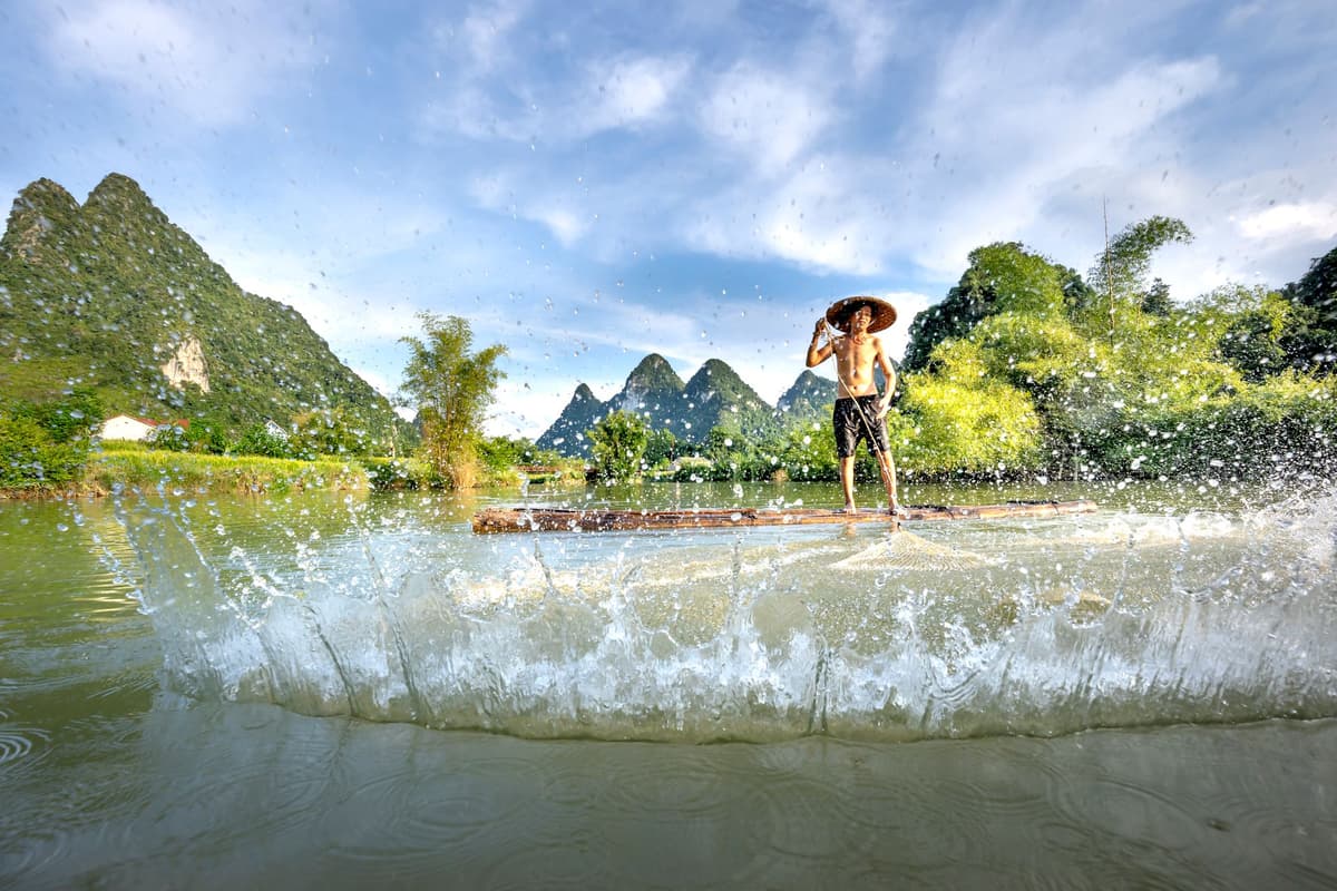 Man fishing with a net in an idyllic Chinese landscape with mountains and water splash.